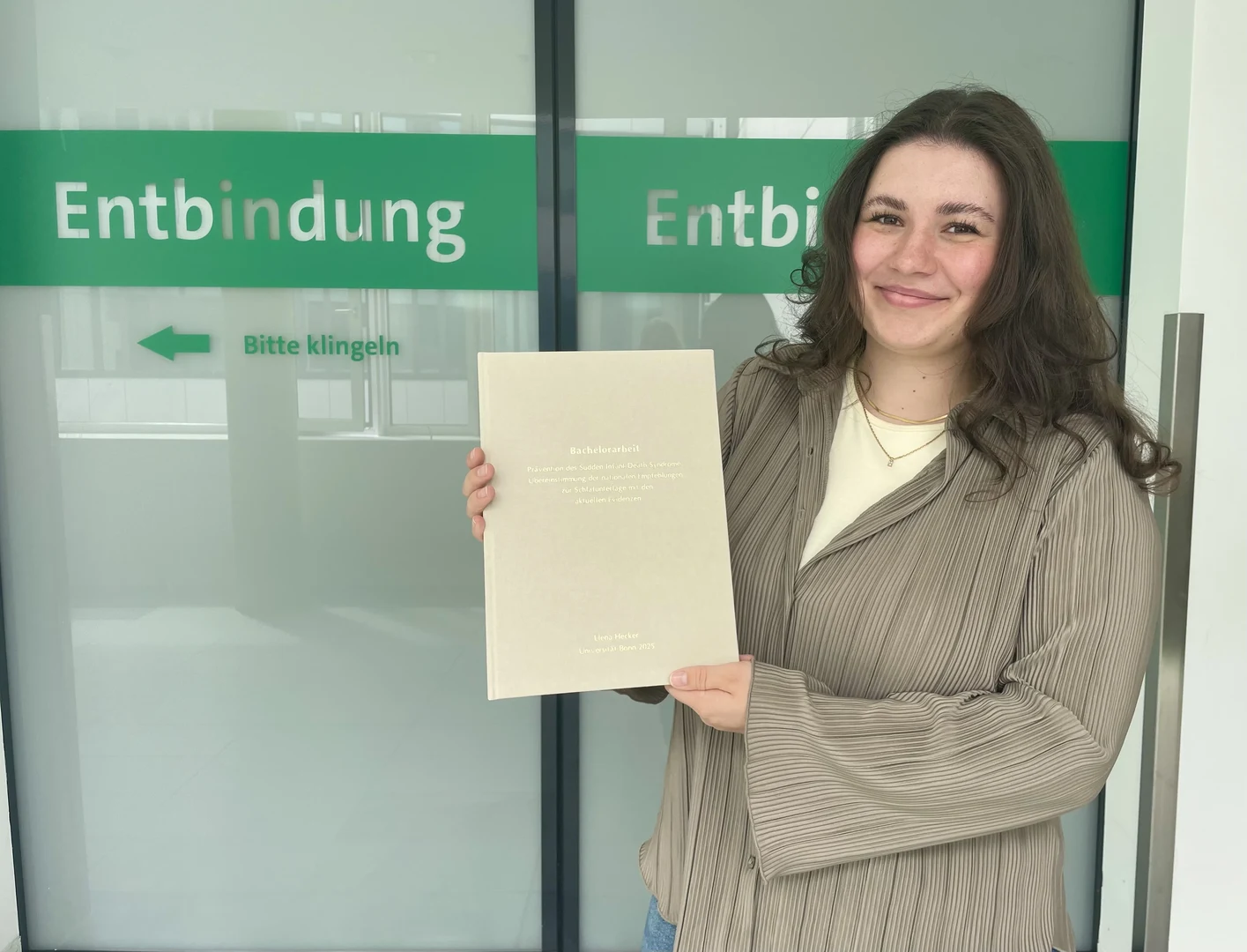 Elena Hecker hat als erste Studierende der Hebammenwissenschaft eine Bachelorarbeit an der Medizinischen Fakultät Bonn eingereicht. Das Foto zeigt sie vor einem Kreißsaal des UKB, in dem sie während ihres Studiums viel Zeit verbracht hat. In den Händen hält sie eine persönliche Druckversion ihrer Arbeit – die offizielle Abgabe erfolgte digital. Foto: Yasmin Kalkan/Medizinische Fakultät Bonn.
