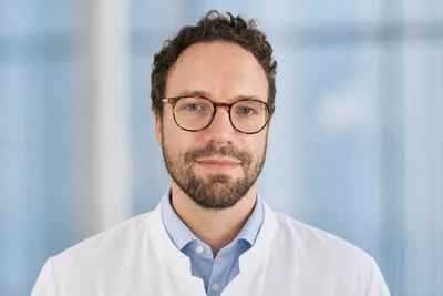Eine Wissenschaftlerin und ein Wissenschaftler arbeiten hinter einer Glasfassade und mischen Chemikalien mit Großgeräten.