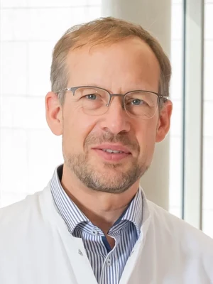 Eine Wissenschaftlerin und ein Wissenschaftler arbeiten hinter einer Glasfassade und mischen Chemikalien mit Großgeräten.