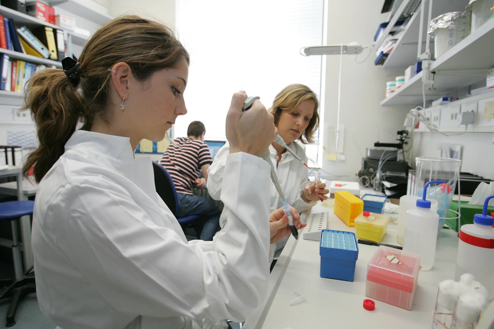 Eine Wissenschaftlerin und ein Wissenschaftler arbeiten hinter einer Glasfassade und mischen Chemikalien mit Großgeräten.